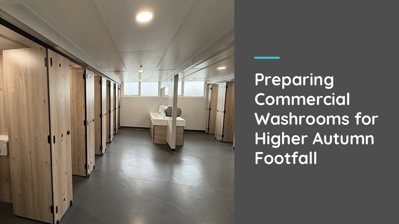 A long, clean commercial washroom with light wood-grain cubicles and a dry grey floor leading to a central basin unit, ready for higher autumn footfall.